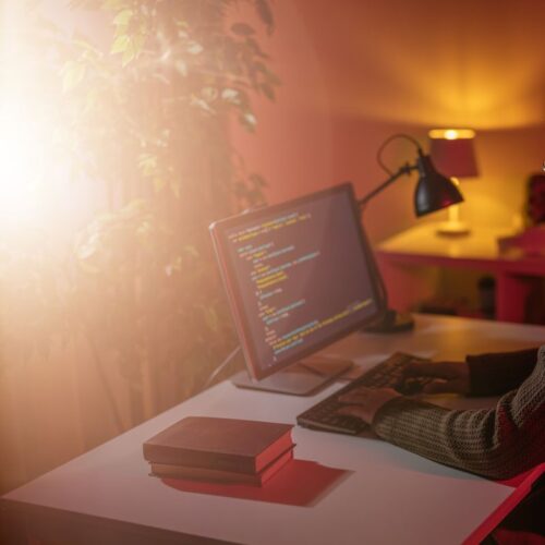 Confident caucasian man in eyewear sitting at desk and programming on modern pc. Orange soft light at dark living room. Remote work at home.